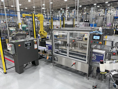 A five-axis robotic arm picks two filled bottles at a time and places them into partitioned corrugated cases. Cases are closed, taped, and receive a label before arriving at the palletizer.