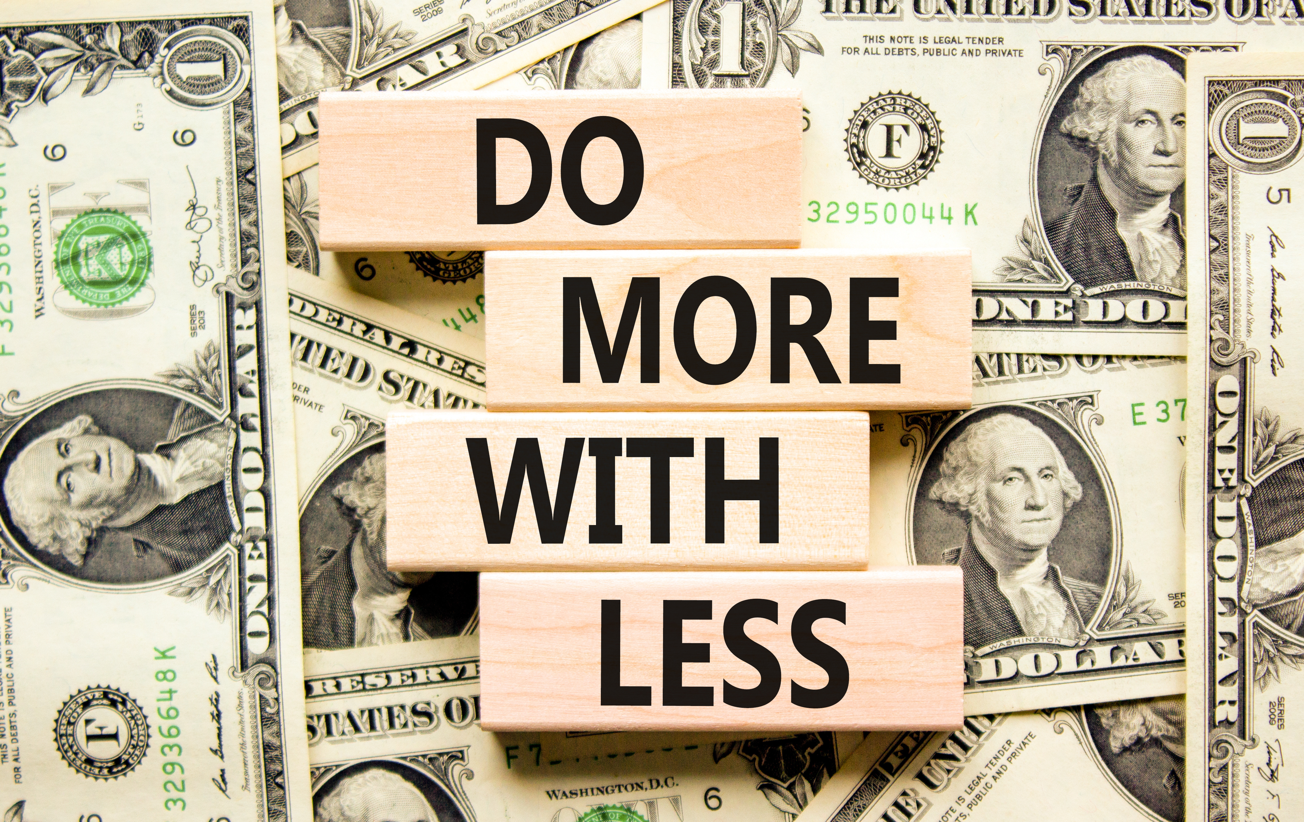 Wooden blocks spelling 'Do More With Less' on scattered US dollar bills