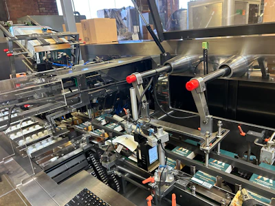 Capsules are pushed from buckets 10 at a time into cartons that are picked from a magazine feed. After cartons are erected and have their bottom flaps closed with adhesive, they’re filled and date coded before being closed and discharged.
