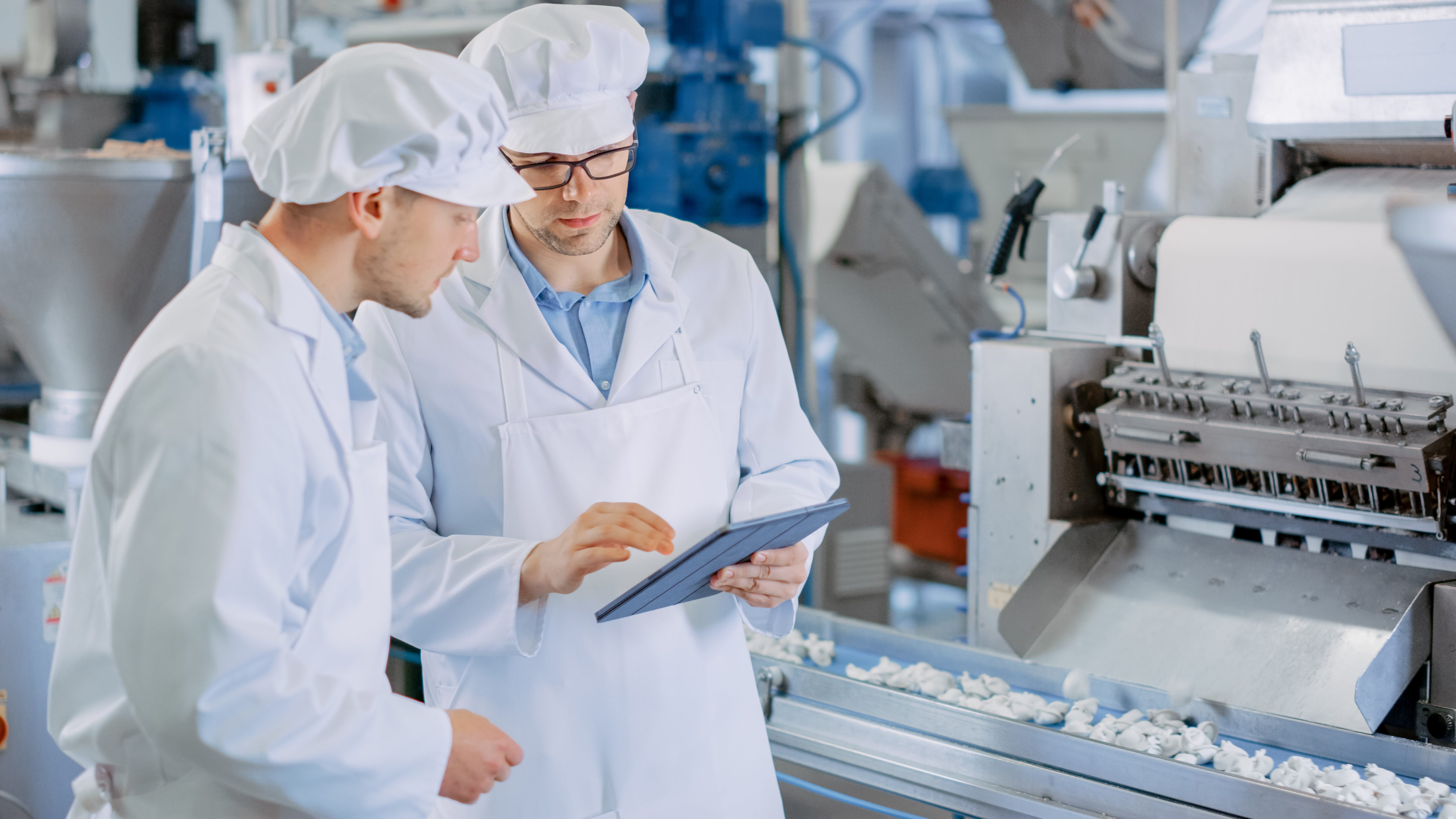 Two workers in white uniforms reviewing data on tablet near food processing machinery