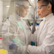 Scientist in white lab coat working with scientific equipment behind glass partition