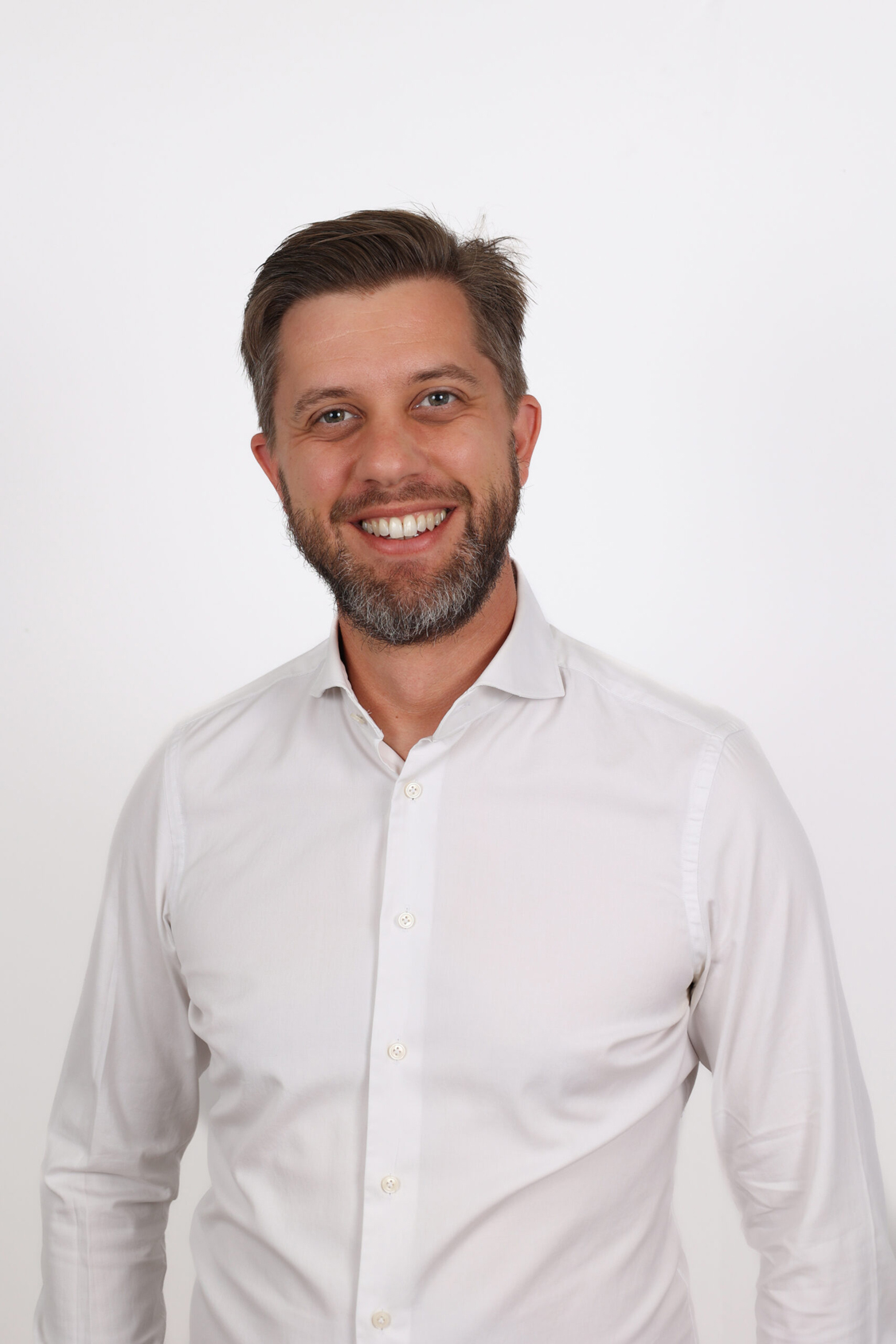 Professional headshot of a smiling man with beard wearing a white button-up shirt against a light gray background