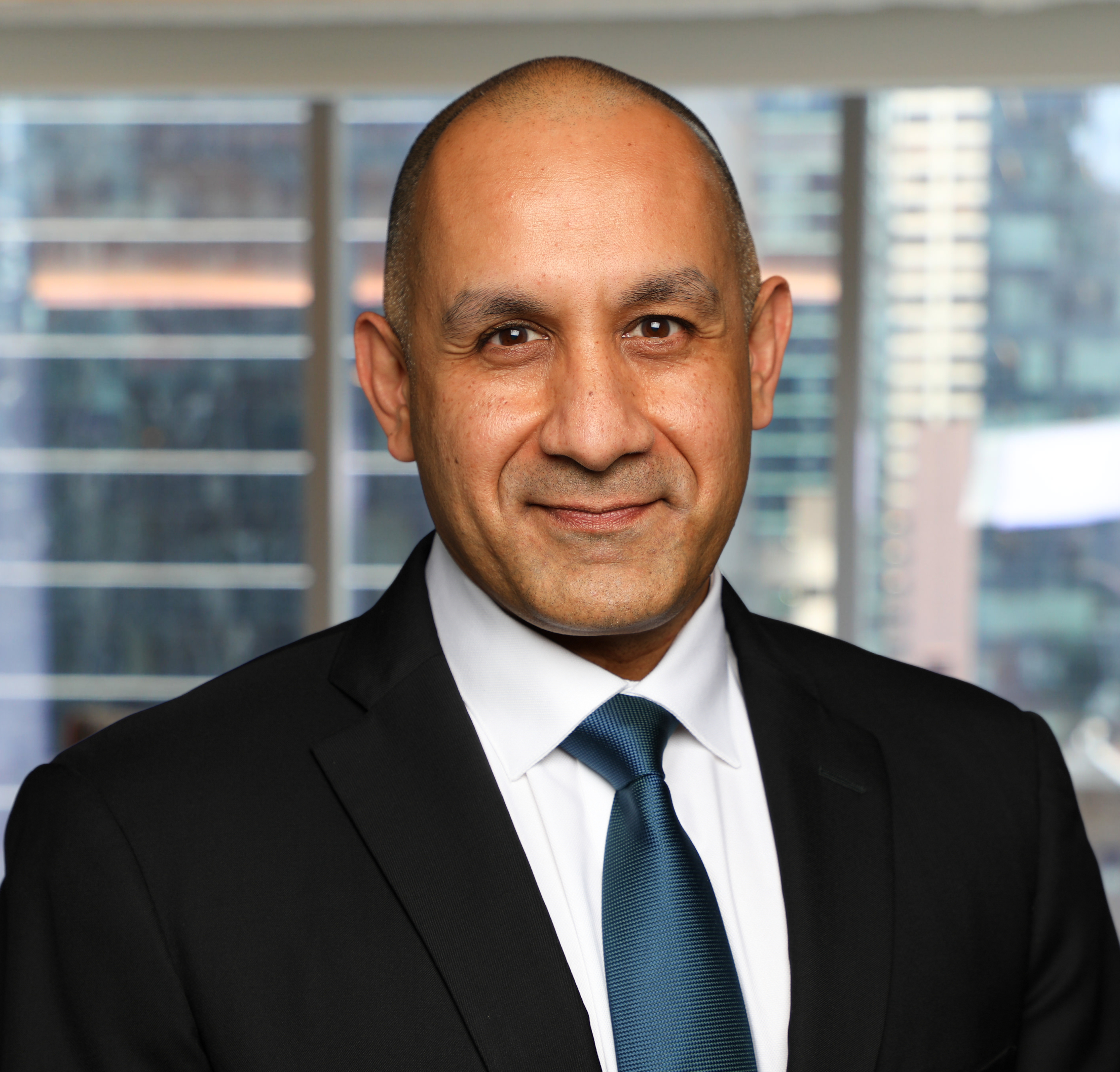 Professional headshot of a man in dark suit and blue tie against office building windows