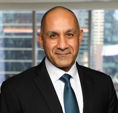 Professional headshot of a man in dark suit and blue tie against office building windows