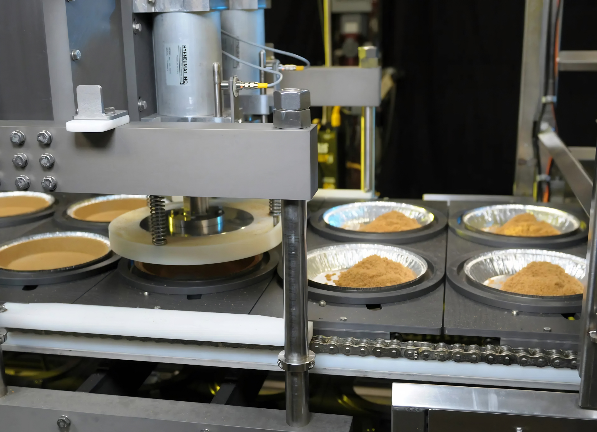 Industrial baking machine with multiple stations dispensing batter into aluminum containers with finished baked goods