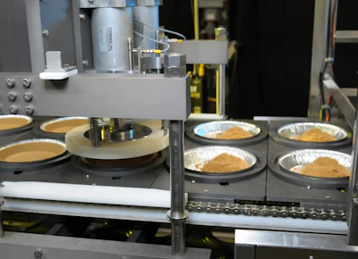 Industrial baking machine with multiple stations dispensing batter into aluminum containers with finished baked goods