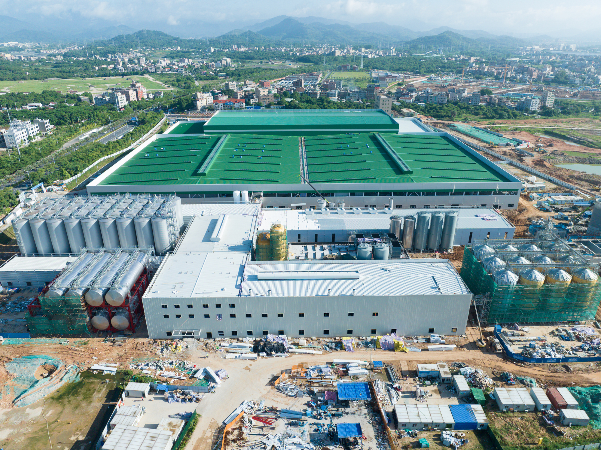 Aerial view of large industrial facility with green rooftop pools under construction, surrounded by urban development and mountains