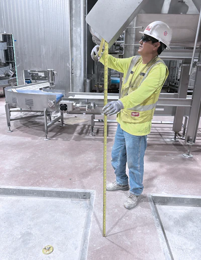 Construction worker in yellow safety gear measures industrial equipment with tape measure on job site