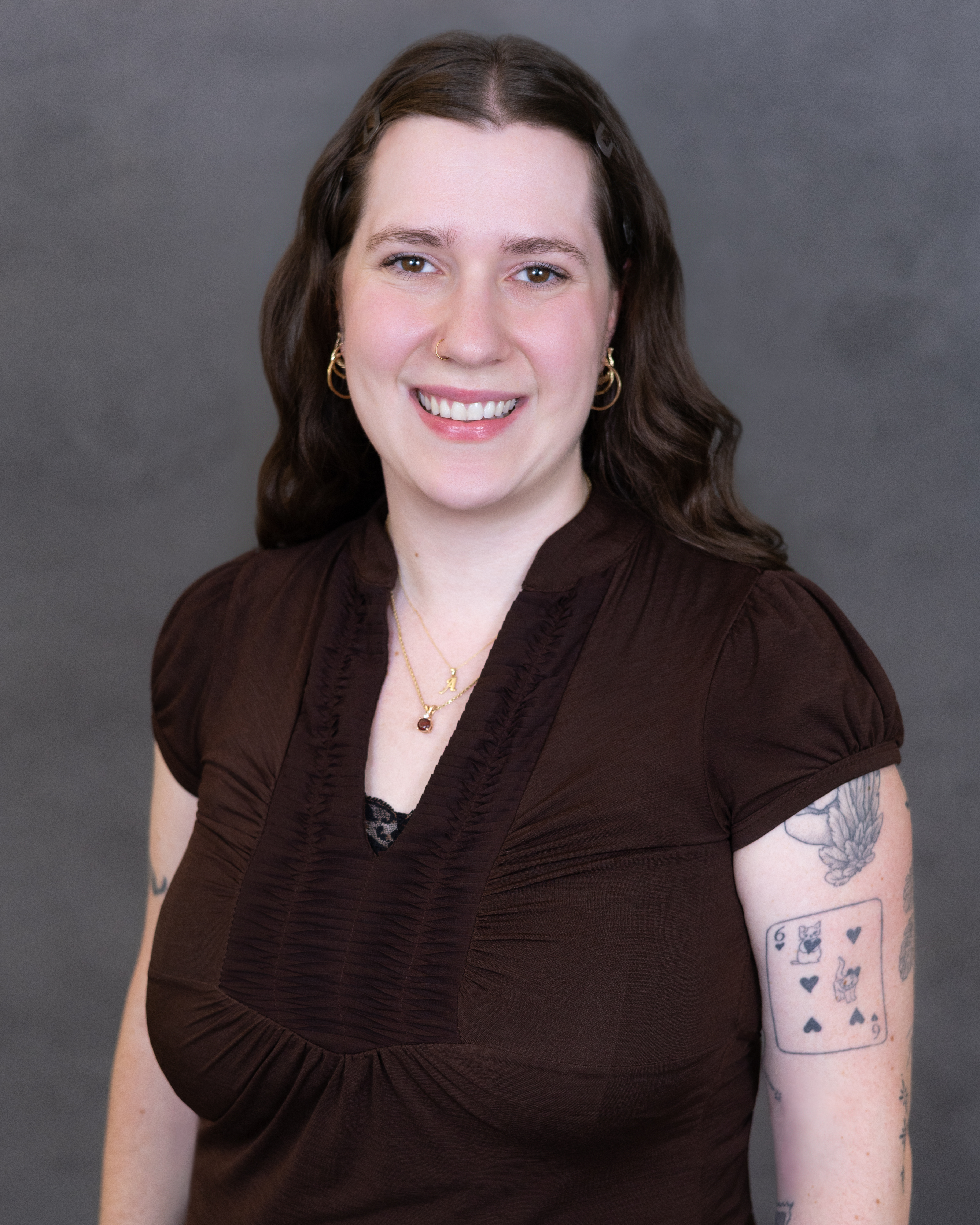 Professional headshot of a smiling woman with dark wavy hair wearing a brown top and gold hoop earrings