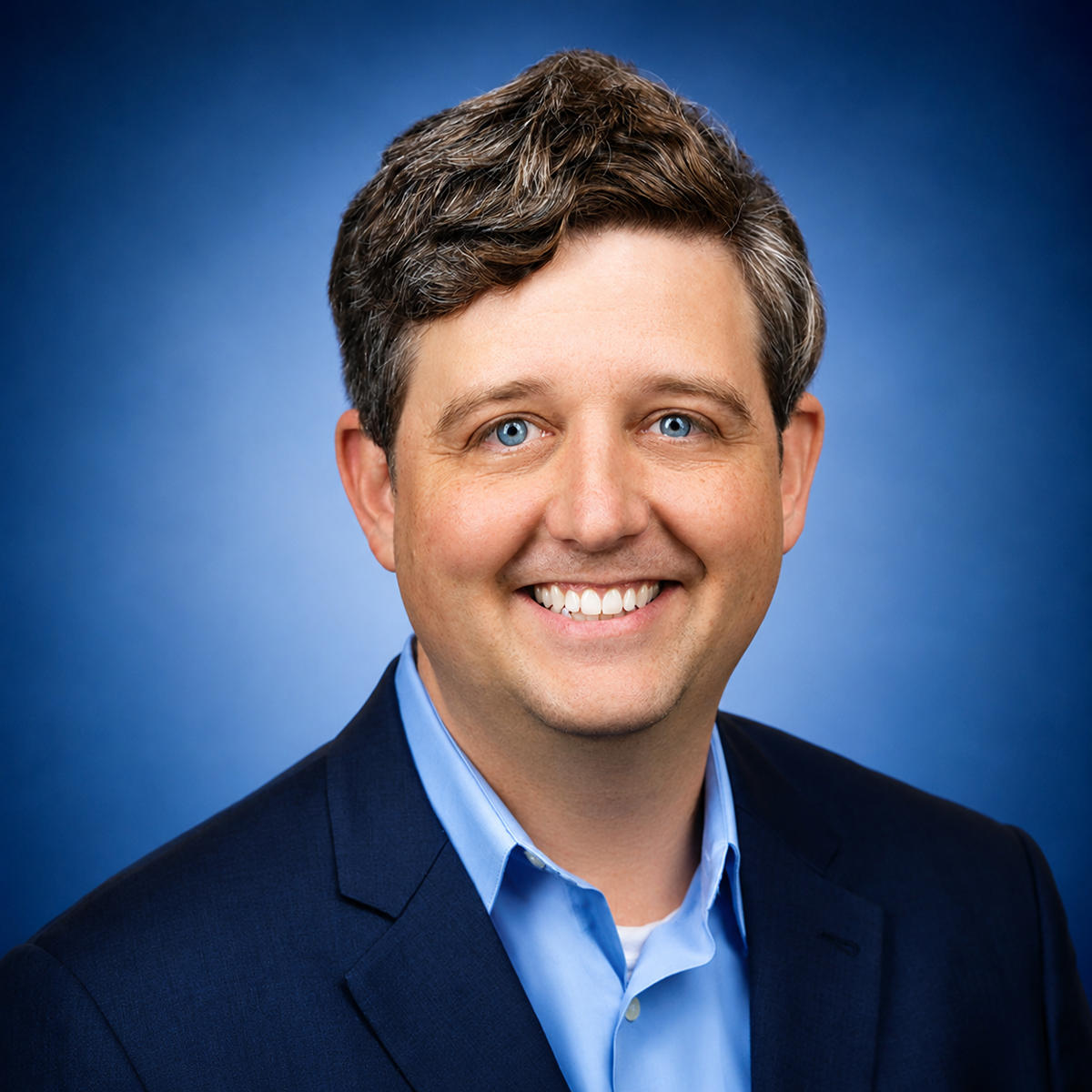 Professional headshot of a smiling man in navy blazer and light blue shirt against blue background