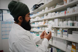 Pharmacist in white coat examining medication bottle in front of pharmacy shelves stocked with pharmaceutical products