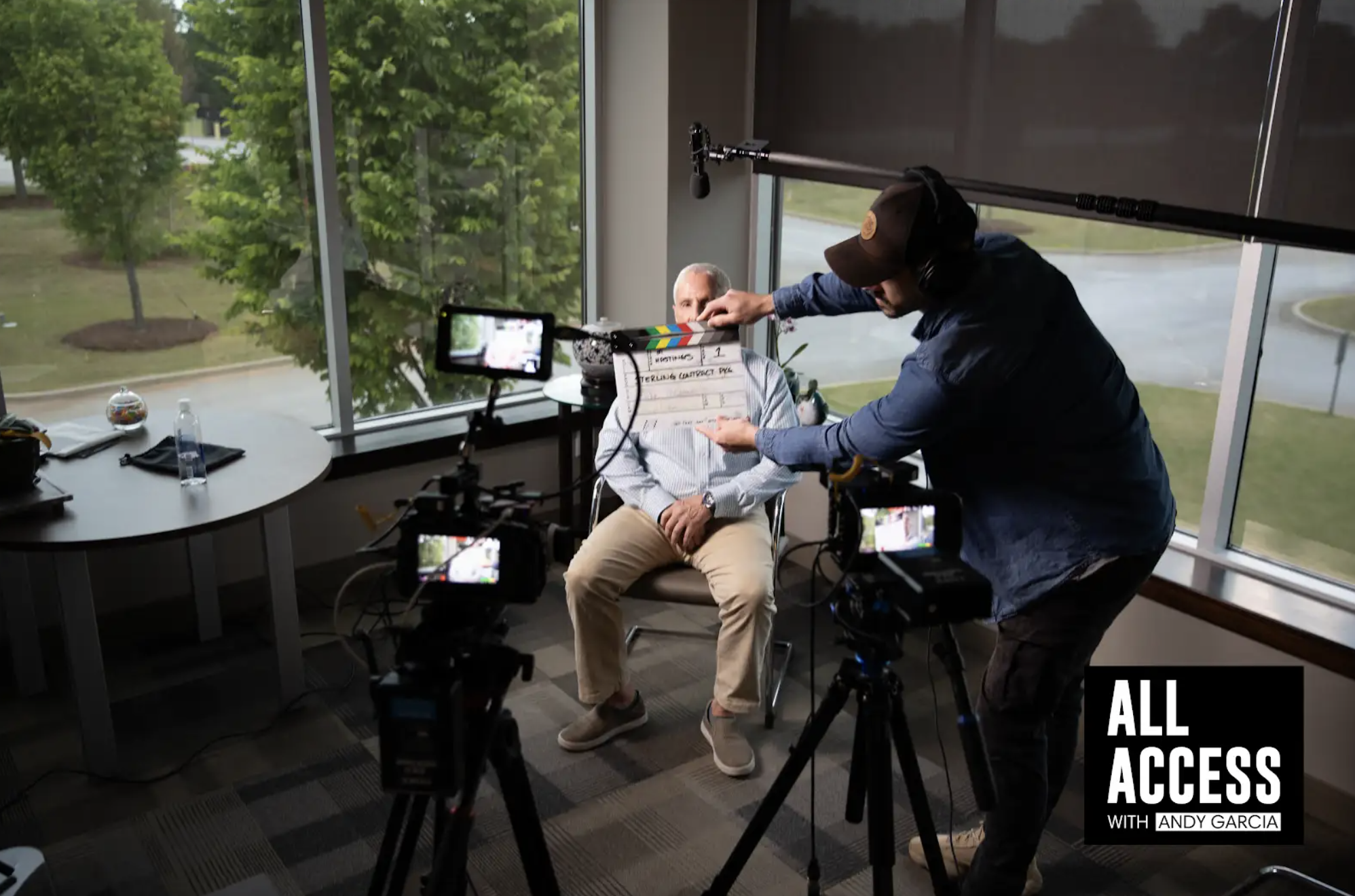 Behind-the-scenes interview filming with camera operator adjusting microphone on subject in modern office with large windows