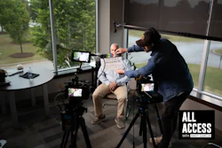 Behind-the-scenes interview filming with camera operator adjusting microphone on subject in modern office with large windows