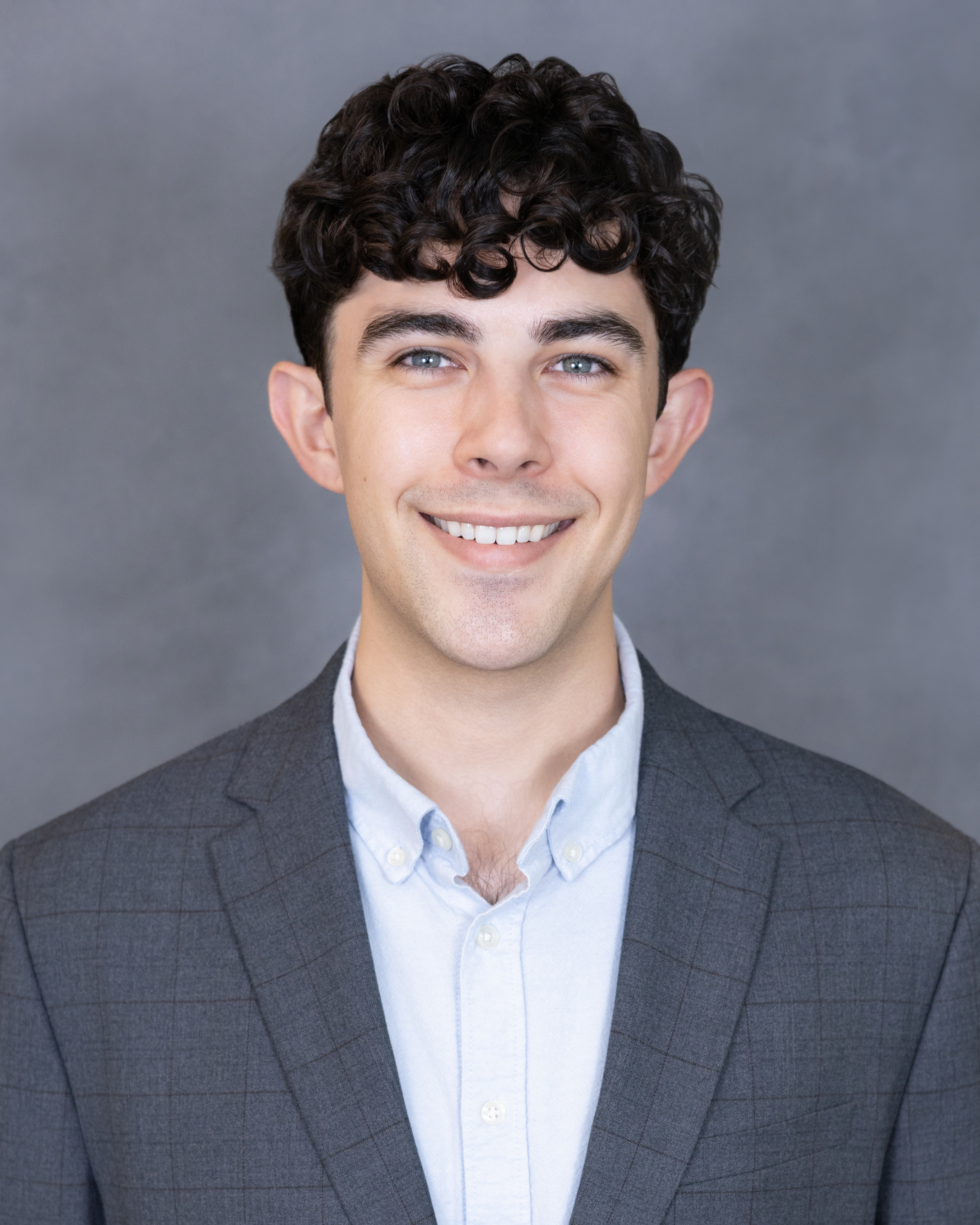Professional headshot of a young man with dark curly hair wearing a gray blazer and light blue shirt, smiling against a gray background