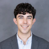 Professional headshot of a young man with dark curly hair wearing a gray blazer and light blue shirt, smiling against a gray background
