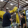 Two technicians service a yellow industrial robotic arm in a warehouse facility with metal framework ceiling.