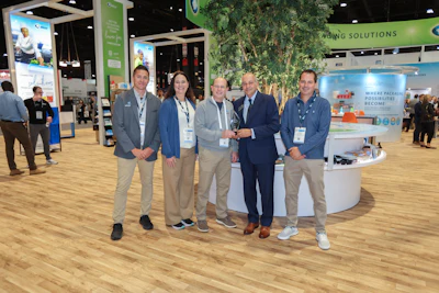 Five professionals in business attire posing at a trade show booth with green branding and wooden flooring